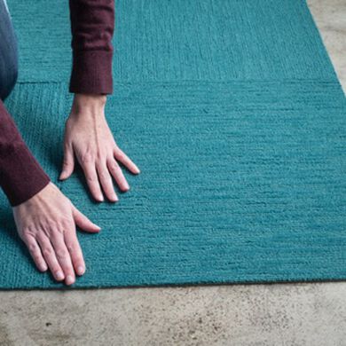 Hand of a person installing teal FLOR carpet tiles on a concrete floor. 