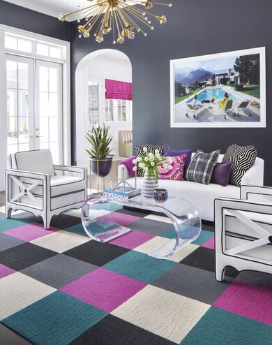 FLOR Made You Look area rug in Bone, Black, Magenta, Slate, and Turquoise, a white couch, purple, pink, and black pillows, white chairs, and a clear table. 