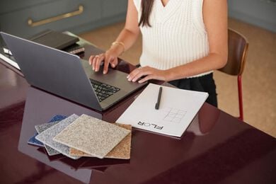 A brunette FLOR area rug Design Consultant in a black jacket  assisting a blonde FLOR customer on a Mac laptop. 