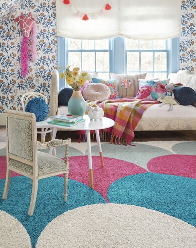 FLOR In the Deep area rug in Bone, Surf, Pink, and Seafoam, a white couch, floral pillows, tartan blanket, and a white table and chair. 