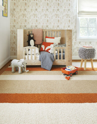 FLOR In the Deep striped nursery rug in Bone, Clementine, and Eggnog, a light wooden toddler bed, and a shaggy gray stool. 