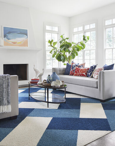 FLOR In the Deep area rug in Bone, Cobalt, and Tidal, a gray couch, blue and orange pillows, and an oval coffee table. 