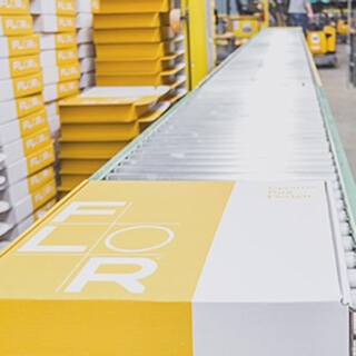 Yellow FLOR carpet tile box on a roller conveyor table in front of stacks of yellow FLOR boxes. 