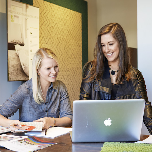 A brunette FLOR area rug Design Consultant in a black jacket  assisting a blonde FLOR customer on a Mac laptop. 